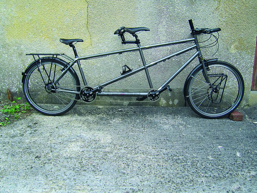 A very dark greeny-silver tandem bike leaning against a wall