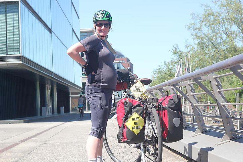 A pregnant woman is standing with her bike. She’s wearing sports gear and a green and black helmet. Her bike is displaying two ‘Baby on board’ signs