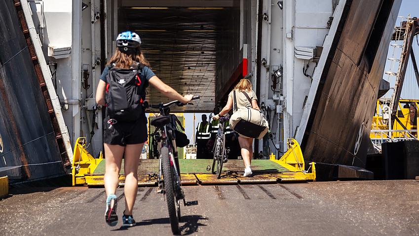 Two cyclists push bikes and carry bags onto ferry on a sunny day