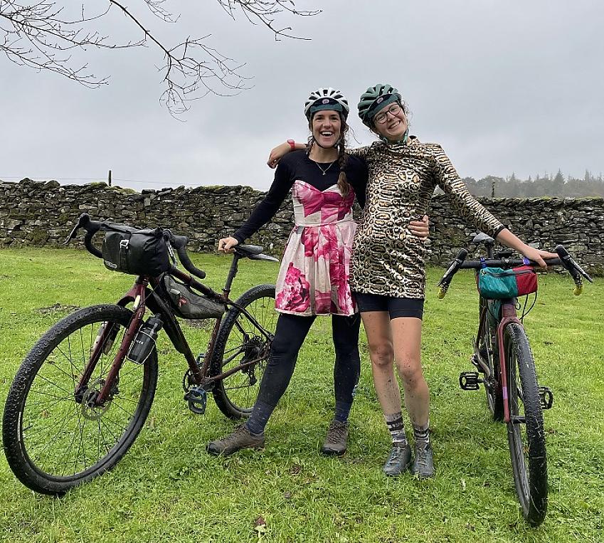 Two women are standing with their gravel bikes. It's a wet day and there's mud and grass on their legs and bikes. The women are both wearing party dresses and are smiling