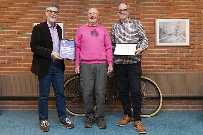 Three men pose in front of a bicycle. They are smiling towards camera. The men to the left and right are holding certificates.