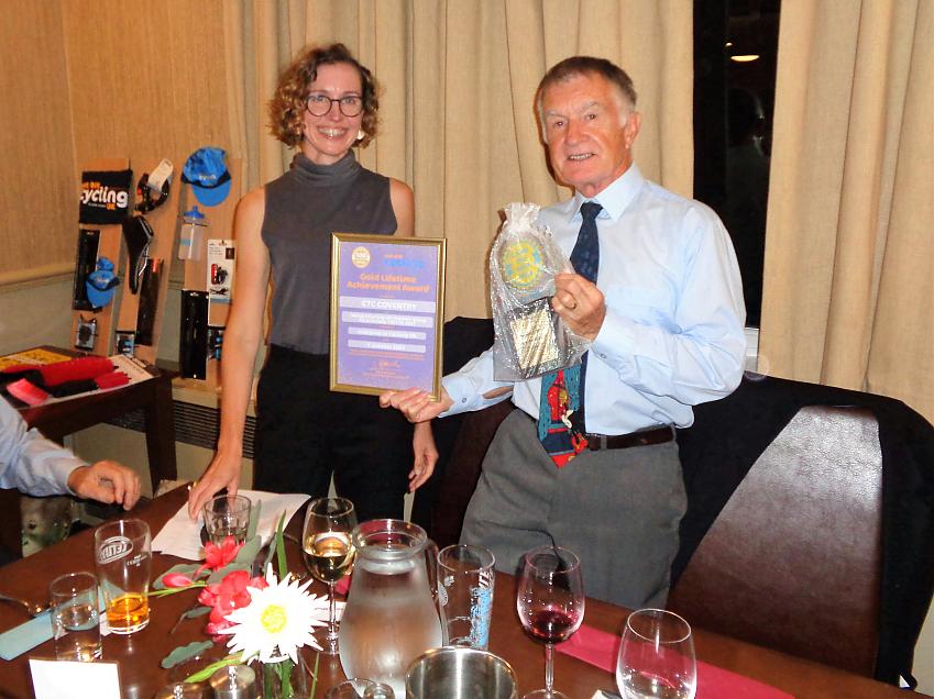 A man and a woman are smiling at the camera. The man is holding a certificate in a frame and a trophy. There are glasses of wine and beer on the table in front of them