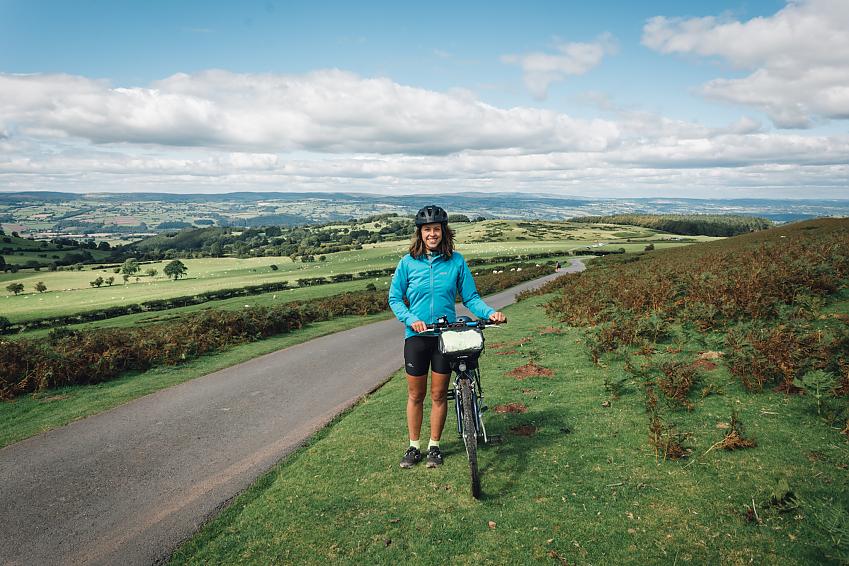 A woman in black cycling shorts and blue jersey stands by the side of a road at the top of a hill.