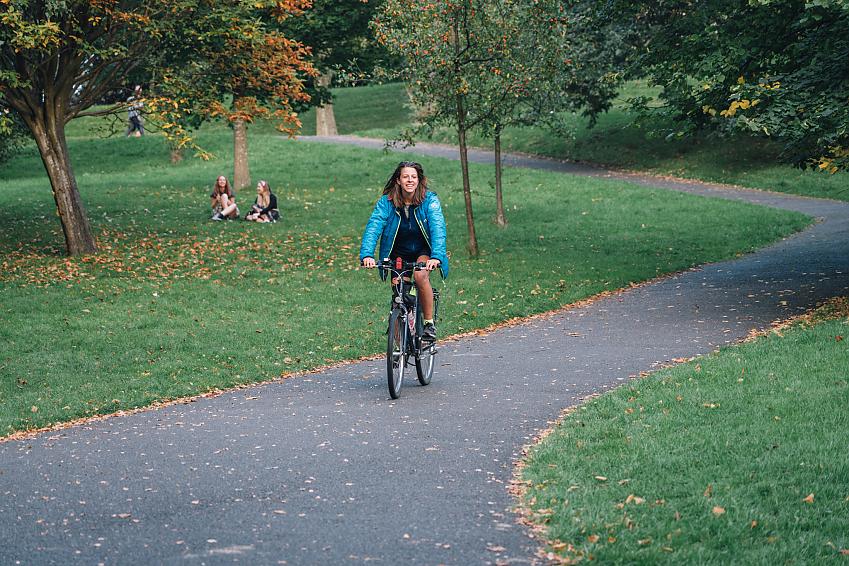 A woman cycling through a park. It's autumn and there are leaves on ground. She is wearing a blue jacket and shorts.