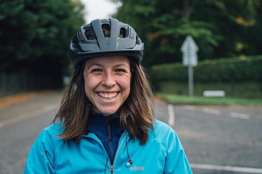 A close-up photo of a woman with long hair wearing a black cycling helmet and blue cycling top; she is smiling