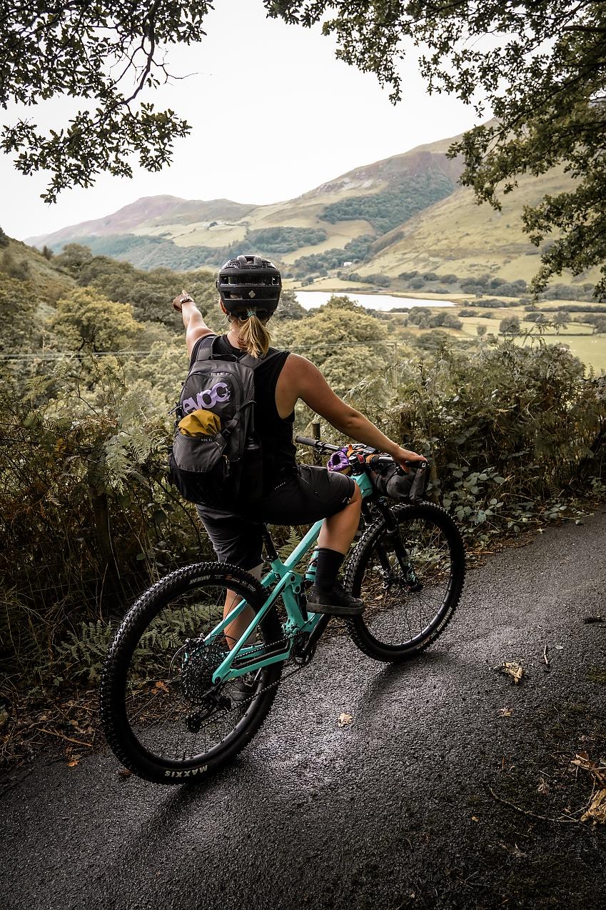 A back view of a woman on a light blue mountain bike pointing towards some hills in the distance. She has a backpack on her back and is wearing cycling shorts and T-shirt and a helmet