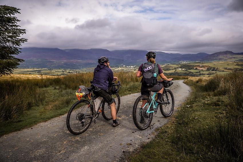 Two people on bikes are stopped on a gravel path. One is a woman on a blue mountain bike, the other is a man on a gravel bike. They are in the mountains