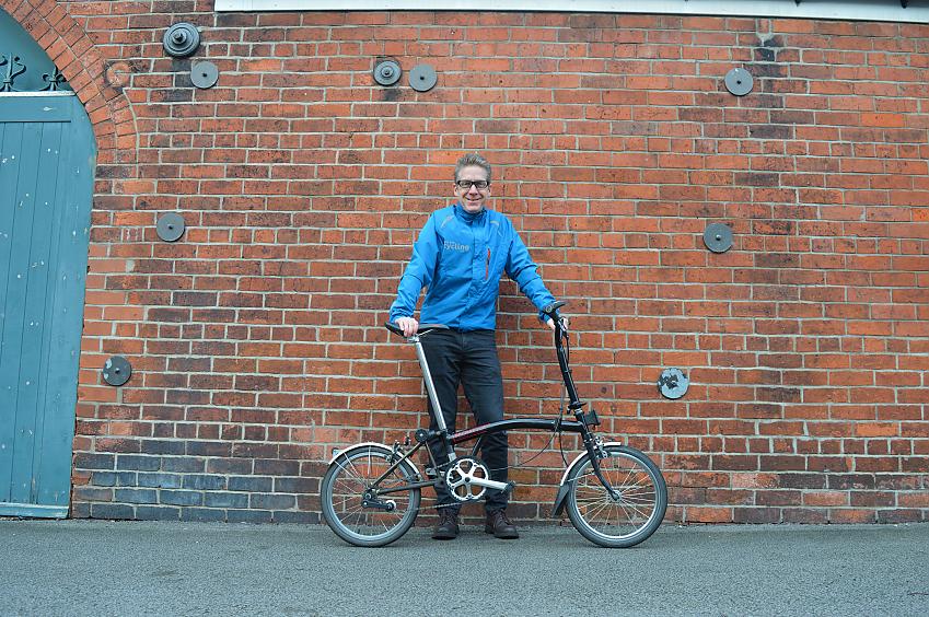 A white man stands against a red brick wall holding a black Brompton foldable bike
