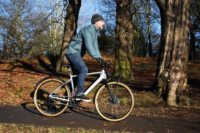 A man wearing a woollen beanie hat, gloves, jacket and jeans rides a silver e-bike through a wooded area on a tarmac path
