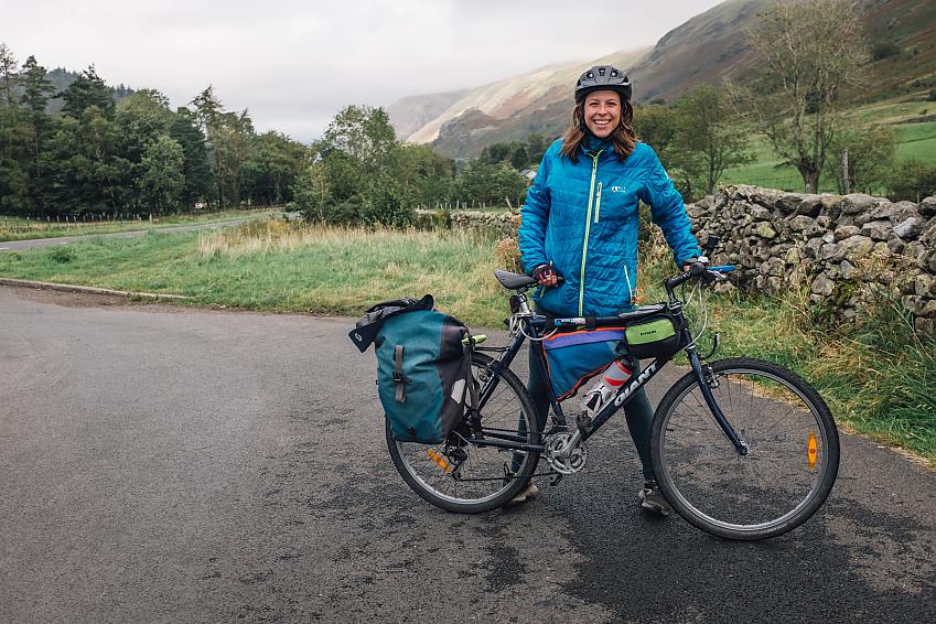 A woman with her loaded-up touring bike. It's raining and she's wearing waterproof clothing. She's smiling.