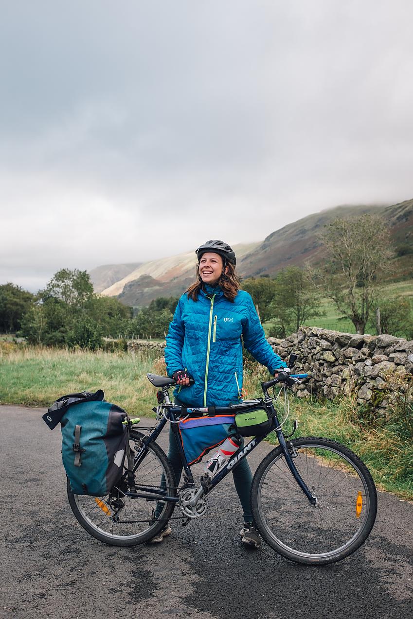 A woman with her loaded-up touring bike. It's raining and she's wearing waterproof clothing. She's smiling.
