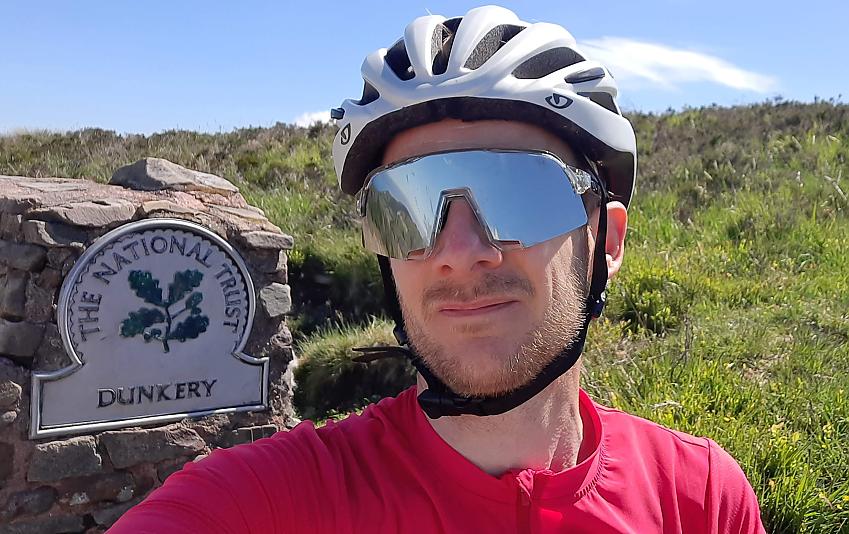 A white bearded man wearing a helmet and cycling sunglasses sits next to a National Trust marker which reads 'Dunkery'