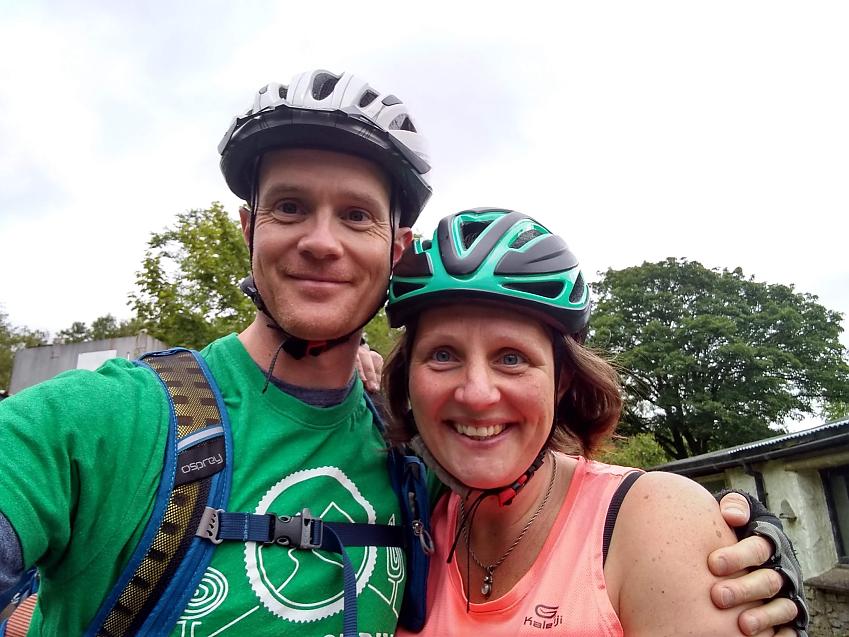 A man and a woman pose together. They are both wearing cycling helmets and clothing. The man’s T-shirt is green, his helmet is white. The woman’s T-shirt is coral, her helmet is green and black. They’re both smiling