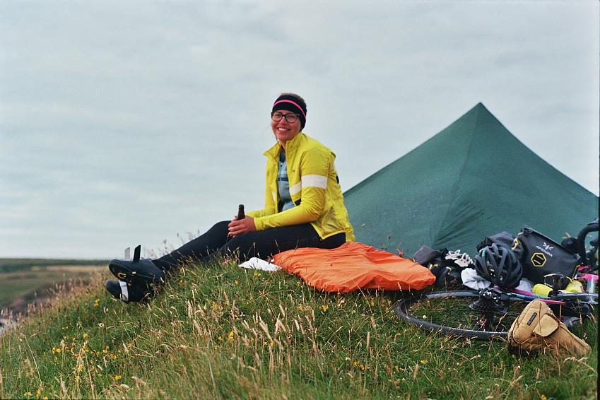 A woman is sitting by her tent. She's wearing a yellow jacket and black leggings and cycling shoes. She's holding a bottle and is smiling. A cycle wheel, helmet and other cycling gear is on the ground next to her.