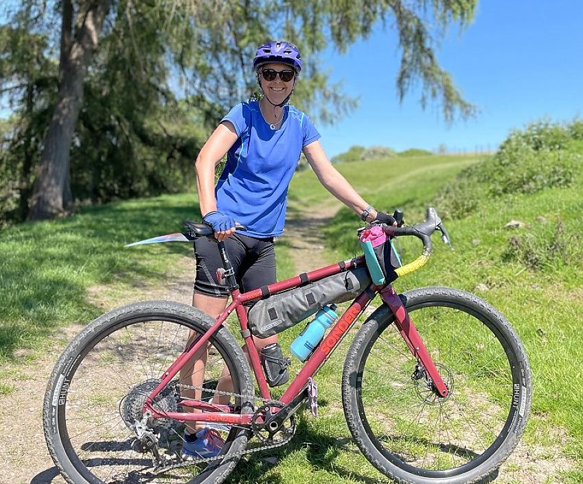 A woman is standing on a dirt path with her red Sonder gravel bike. She is wearing black cycling shorts and a blue jersey. It's sunny and she is smiling