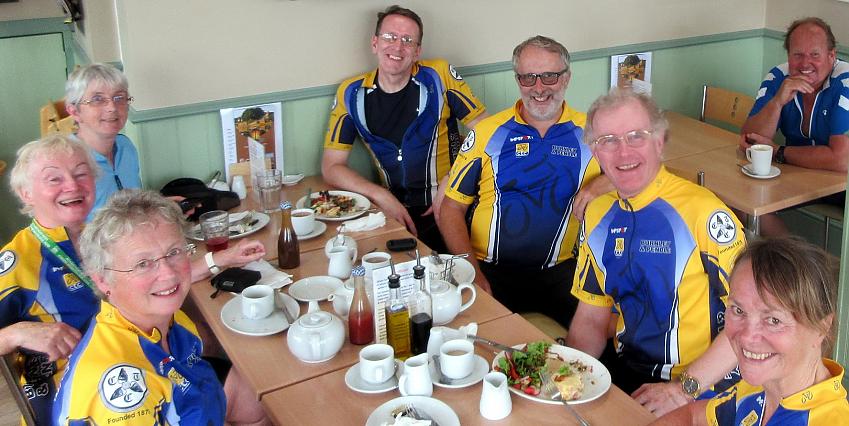A group of cyclists all wearing blue and yellow club kit is sitting at a table enjoying a well-deserved lunch