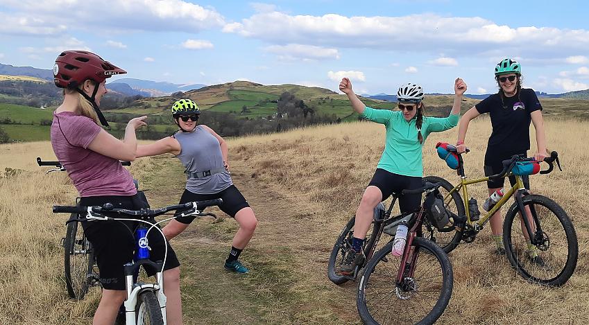 Four women with their bikes at the top of a hill. They look like they're messing around - pulling funny poses for the camera