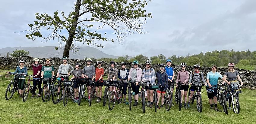 A large group of women and non-binary people are lined up with their bikes in the countryside