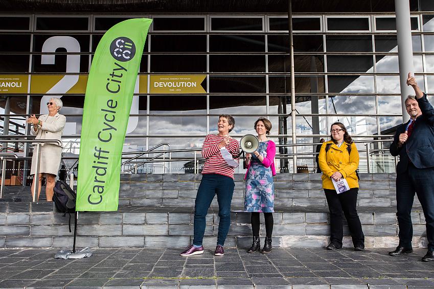 Four women and a man are pictured outside the Senedd in Wales. They are there for an event and one of the women is holding a loud speaker. The man is giving a thumbs up and one of the women is clapping