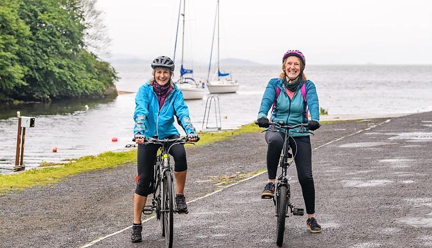 Two women with bikes standing in front of a lake with two boats in the background. The women are both smiling; they are wearing black leggings and blue tops.