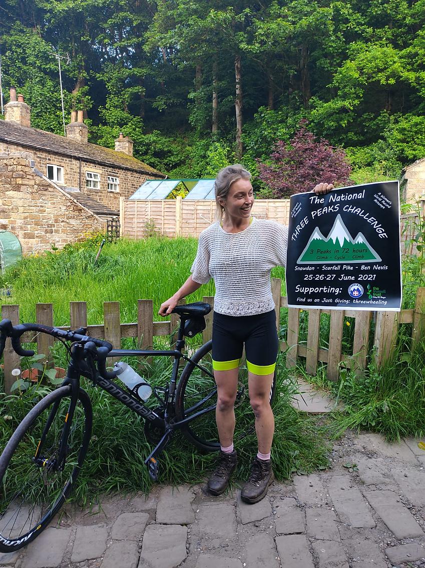 Emily Cowper-Coles posing with her bike and the fundraising campaign poster, a cottage and a woodland in the background