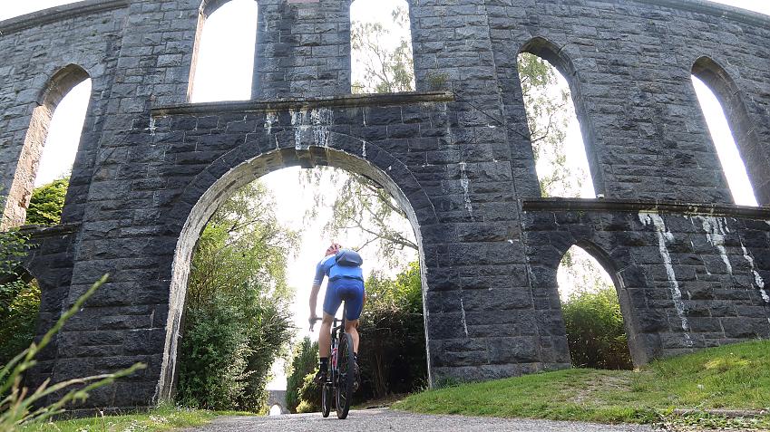 A man cycles towards a viaduct-type structure which has door and archways built into it. A gravel track runs through the centre