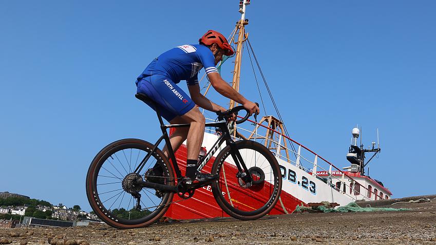 A man cycles in full lycra on an off-road bike in front of a fishing boat
