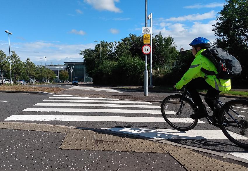 A boy is cycling to school. He's cycling over a zebra crossing. He's wearing a high-vis jacket and helmet. His school bag is on his back