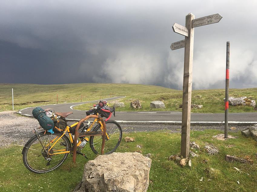 A fully-loaded bicycle rests against a sign post next to a deserted road. A dark rain cloud approaches