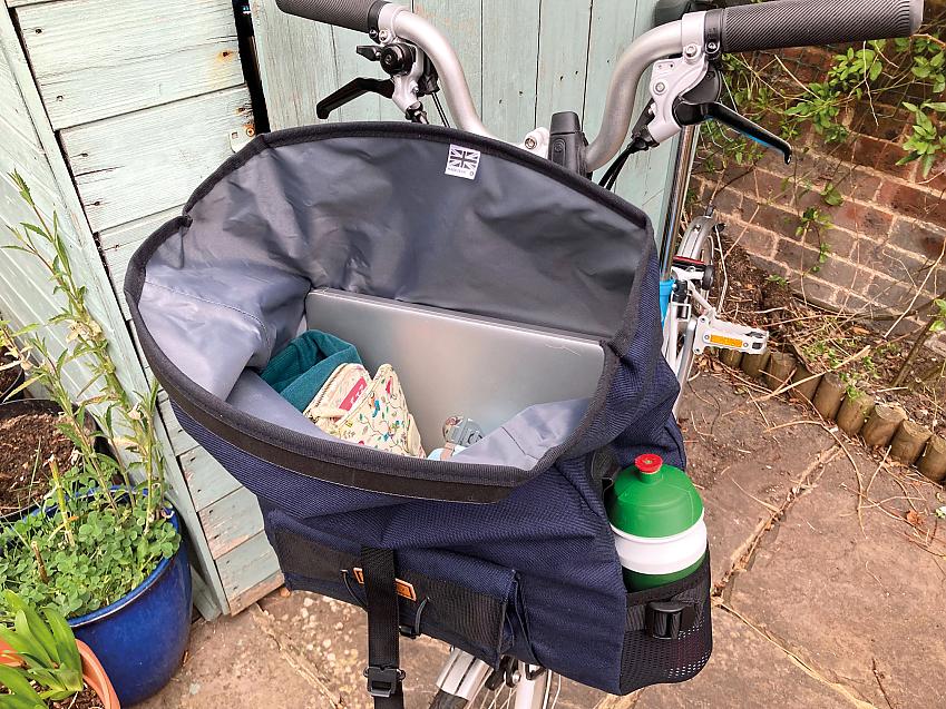 An open bag is attached to the front of a Brompton bike, which is leaning against a small shed in a garden. The bag has a laptop and notebook in it and there's a water bottle in a side pocket