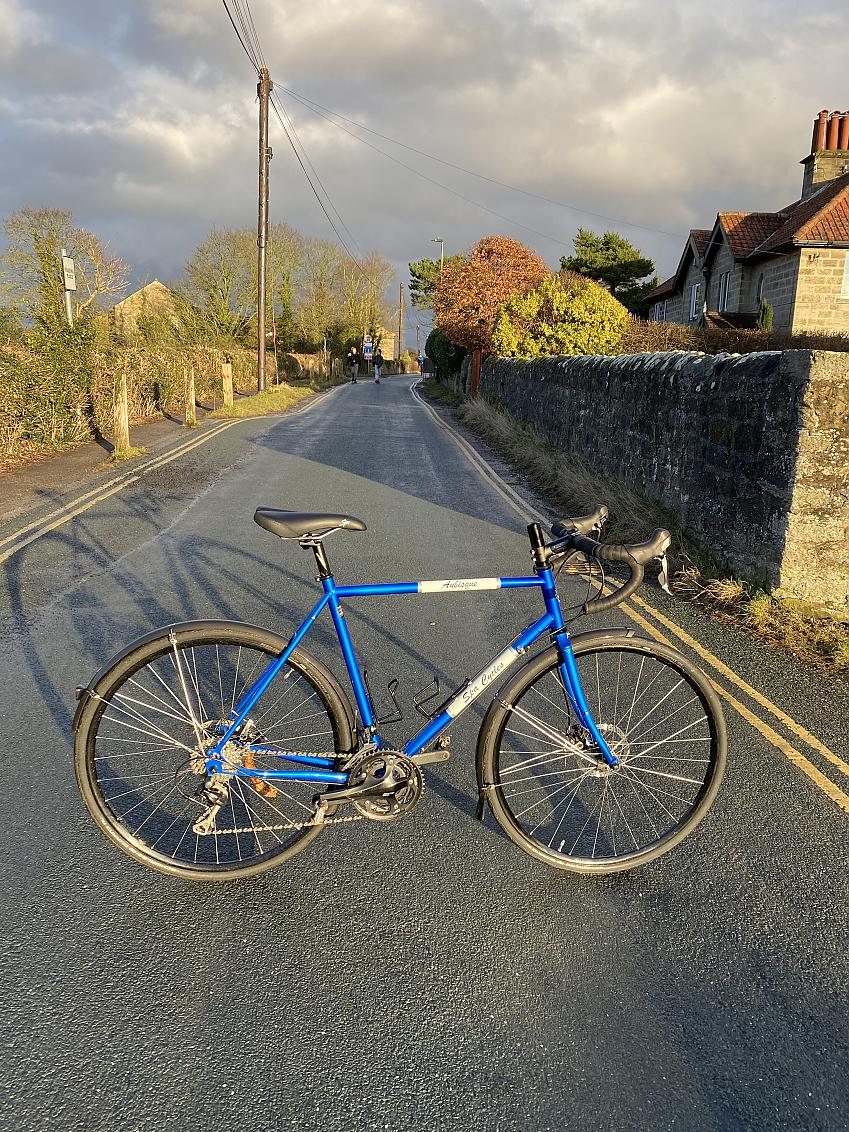 A blue road bike is propped up in a quiet village road with a couple of people walking in the background