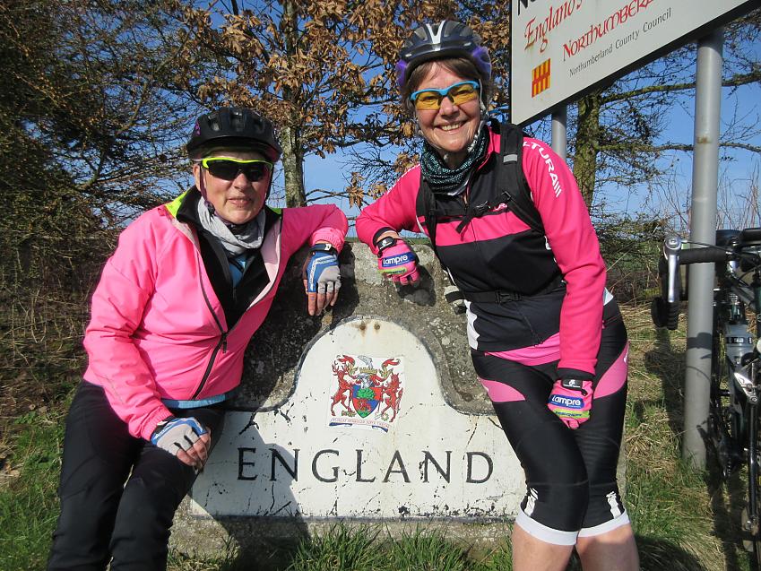 Two women in pink and black cycling kit are sitting on a sign that reads England