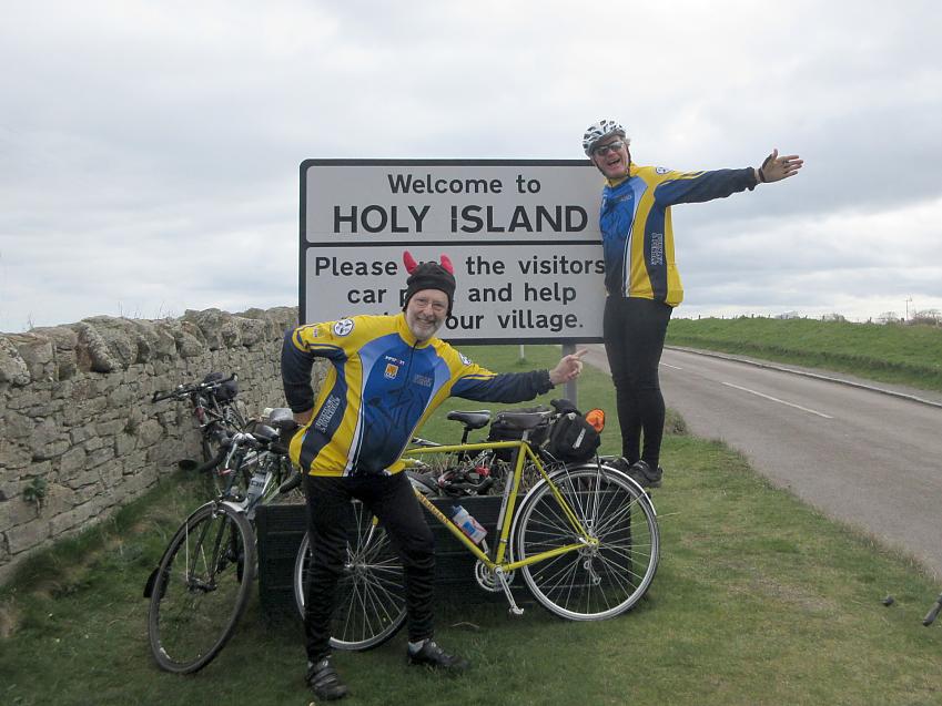 Two male cyclists wearing blue and yellow club kit are posing next to a sign reading Holy Island. They both have touring bikes