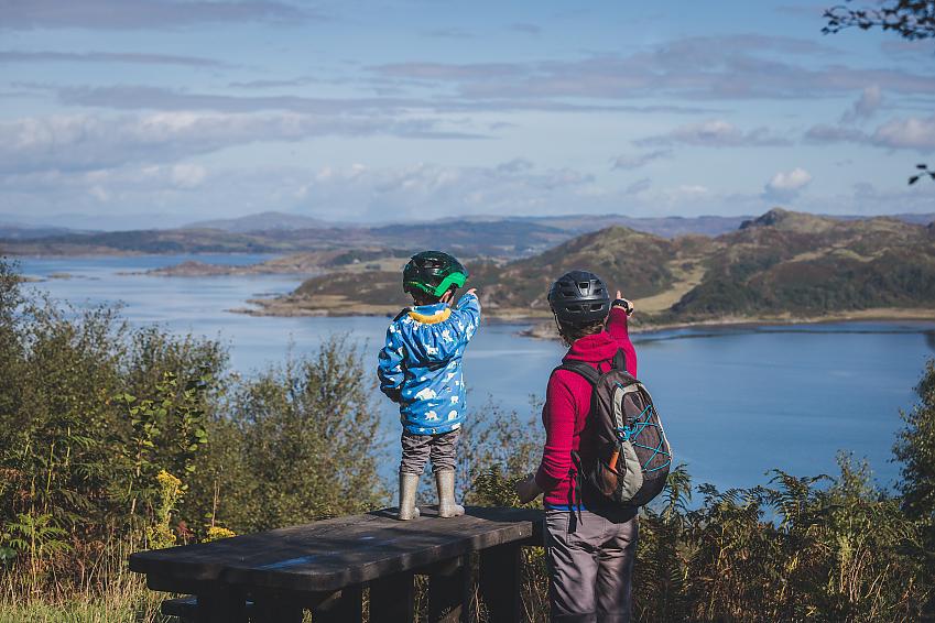 A young boy stands on a bench, his mother stands next to him on the ground. They both point and look at a group of hills across a lake