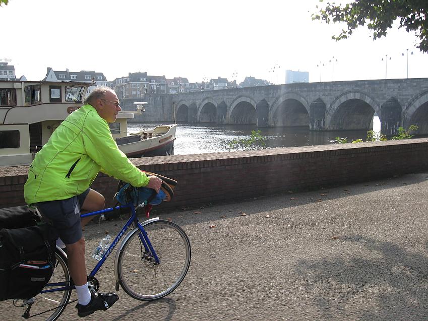 A man cycles along a cobbled tow-path next to a river. In the background a large stone bridge crosses the river