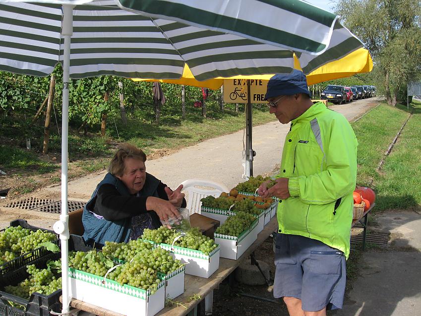 A man stands by a market stall where the owner is selling grapes. Punnets of green grapes cover the table