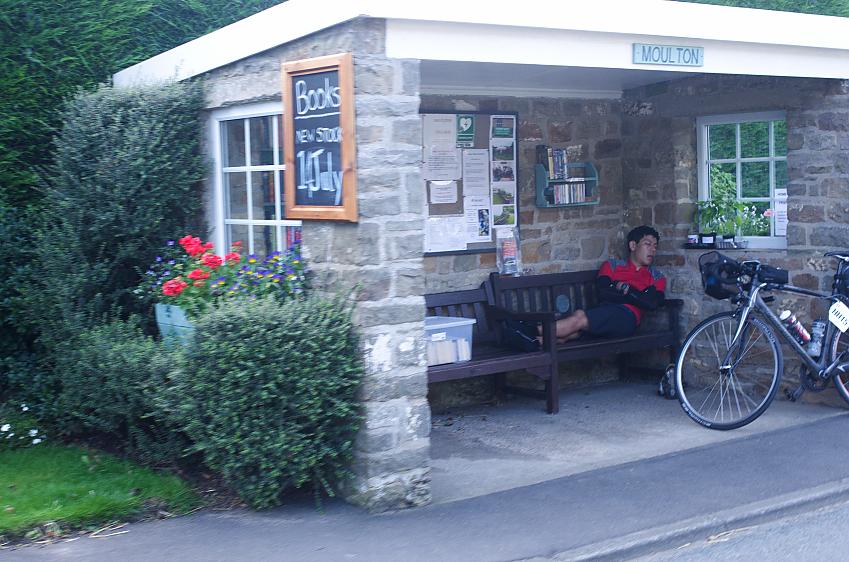 A tired rider sleeps on a bench under an outdoor shelter with information on the local area and shelves of books that are being sold