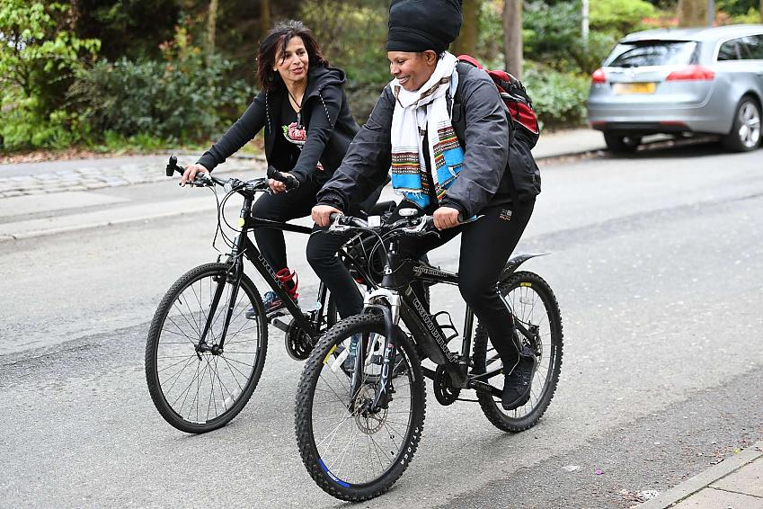 Two women are cycling along a road. They are wearing normal clothes; one has a turban and backpack. They are smiling and chatting