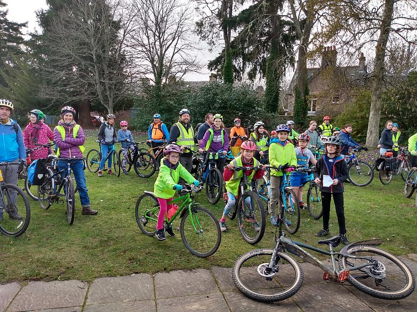 A big group of children with a few adults are all gathered together with their bikes ready to set off on a bike ride. They are of various ages, with all kinds of bike