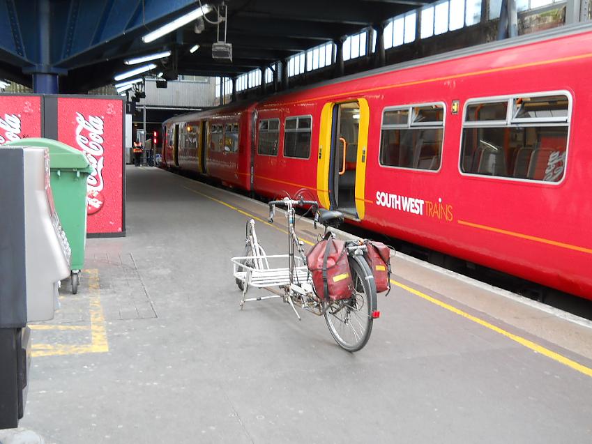 A cargo bike stands on a train platform in front of a train. The cargo bike has panniers on the back, and a large flat-bed area for transporting goods