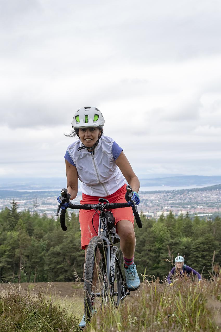 A woman is riding a gravel bike over rough ground and uphill. The bike is black. She is wearing orange shorts, a blue T-shirt, white gilet and helmet and blue shoes. She looks determined but happy