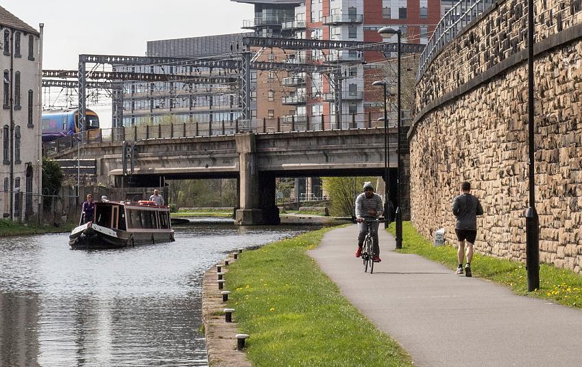People cycle and run along a towpath in an industrial area as a barge passes on the canal