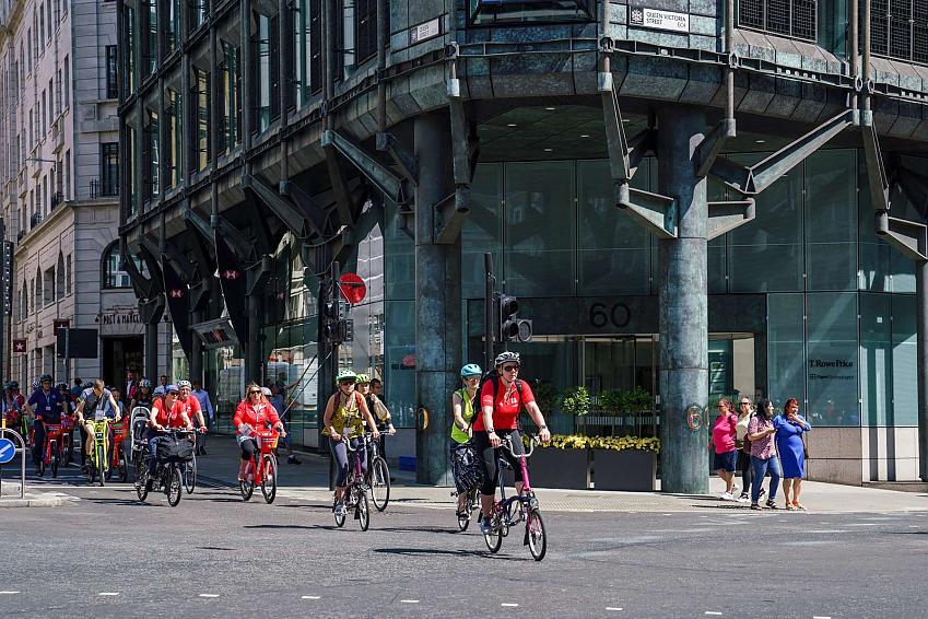 A large number of people are cycling across a junction in London. A big, glass office building is in the background.