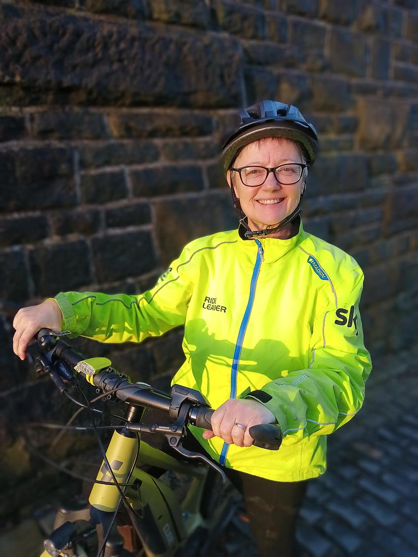 Kate Wylie with her bike, wearing a bright yellow hi-vis jacket and a helmet, smiling at the camera