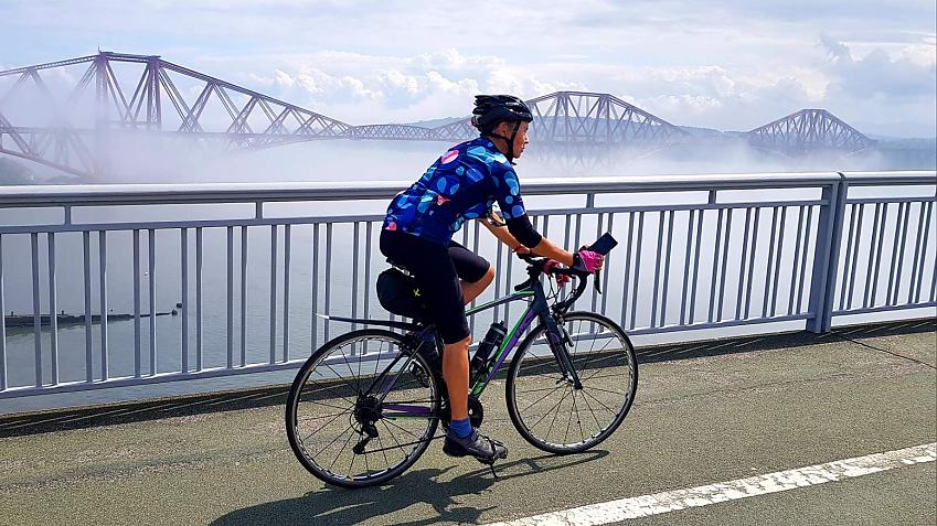 A woman is riding across a bridge. She is wearing cycling gear and riding a Liv road bike. The Forth rail bridge is in the background and it's quite misty