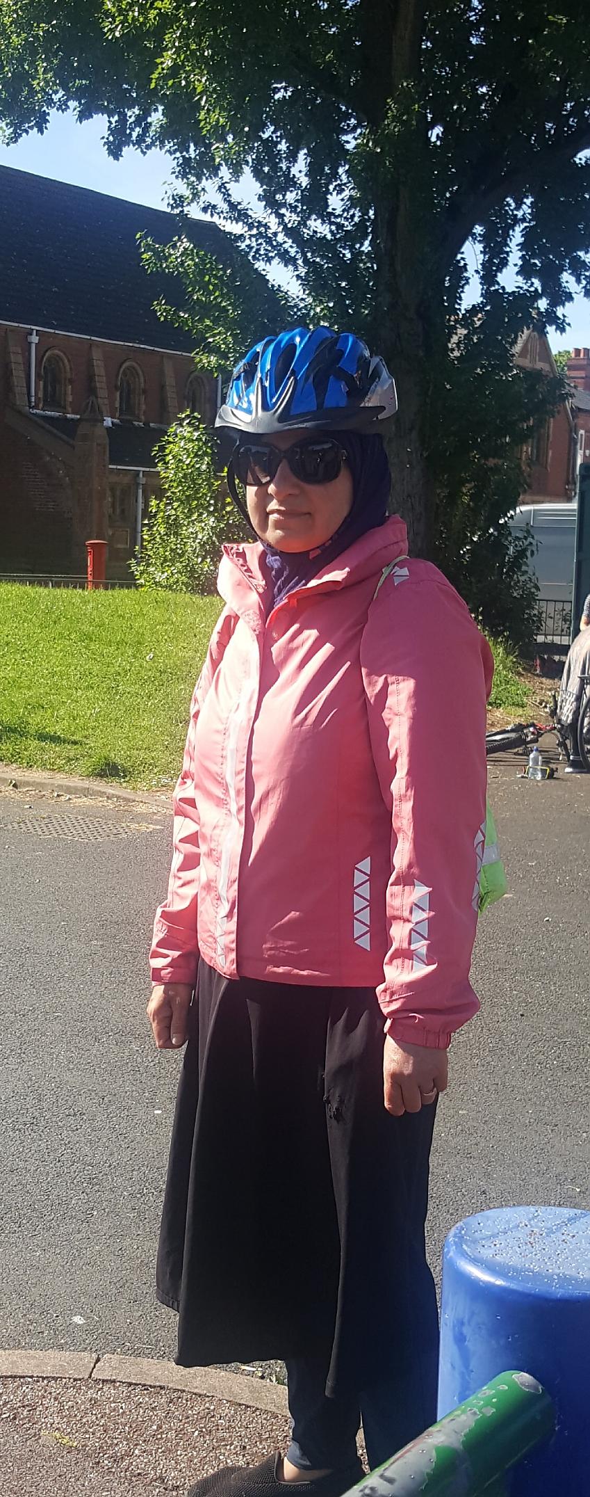 A woman is standing on the pavement. She is wearing a blue cycling helmet, sunglasses, a bright pink hi-viz jacket and a headscarf.