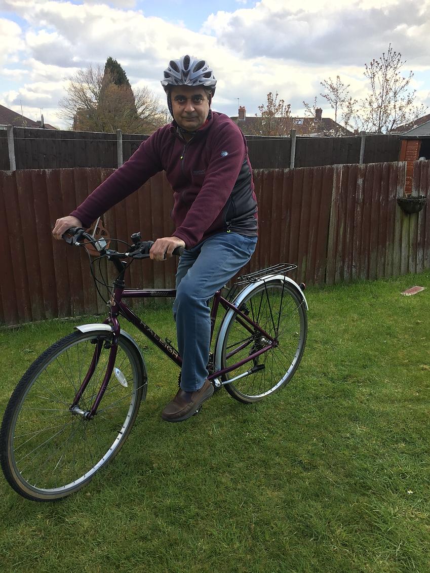 A man stands over his bicycle in a garden. He is wearing casual clothes and a helmet