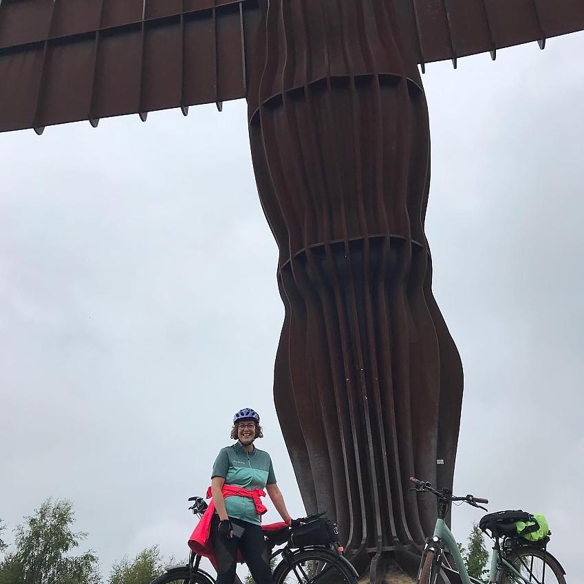 A woman poses with her bicycle in front of the Angel of the North, a giant and imposing sculpture. She is smiling and wearing cycling clothes