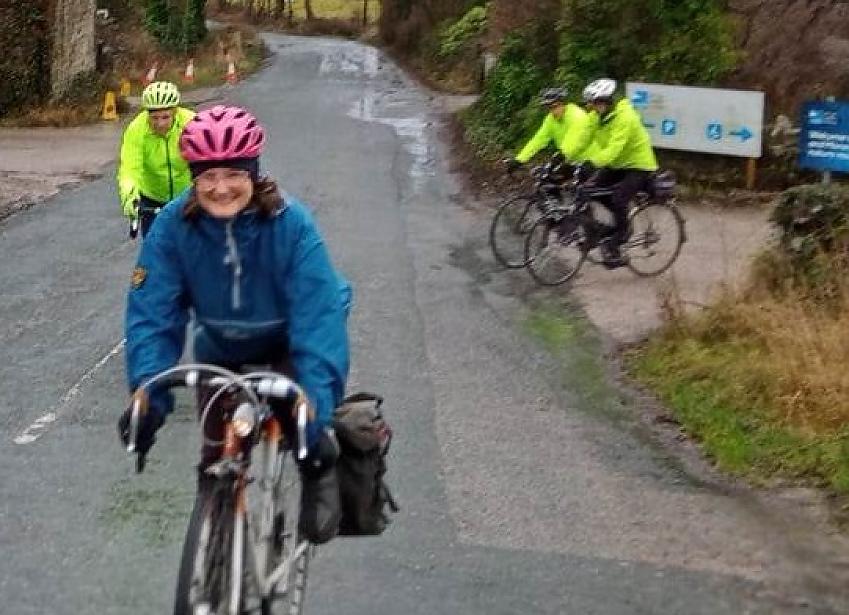 Four cyclists ride towards us on a quiet country lane, it's raining and they are wearing waterproofs