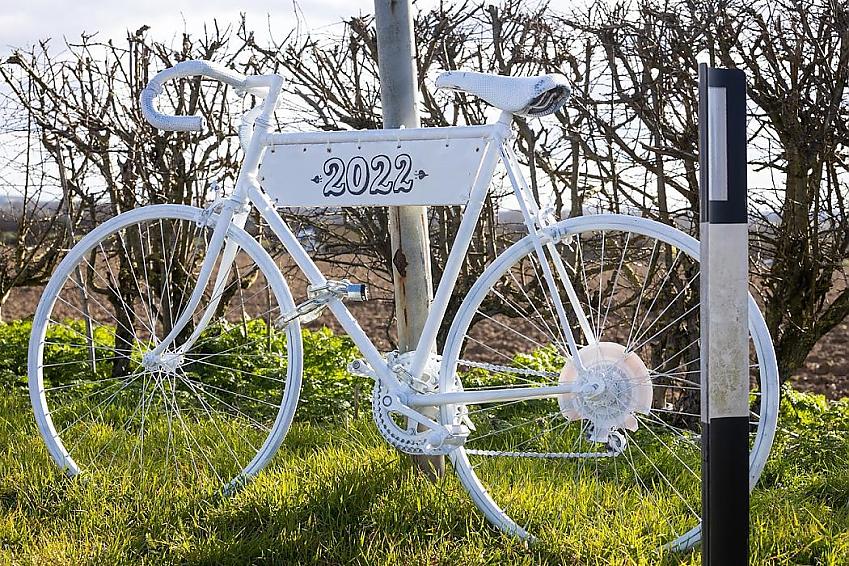 A white-painted bike is placed, chained to a lamppost at a road junction. Under the bicycle top-tube a white banner has been created, showing the year '2022'